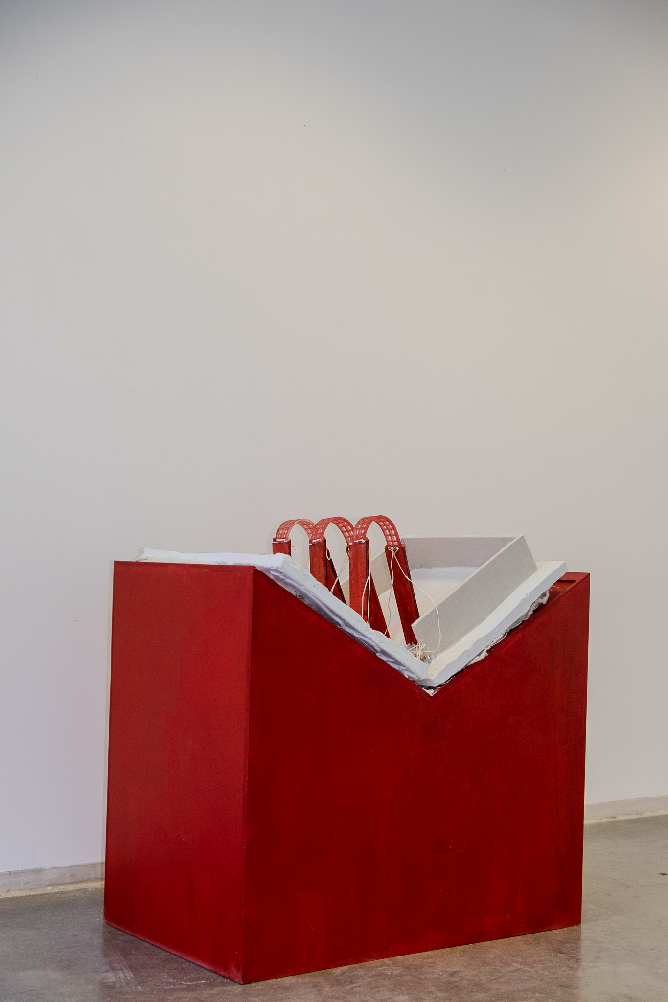 A red geometric sculpture with white and gray slabs forming a book protruding from the top, displayed against a plain white wall.